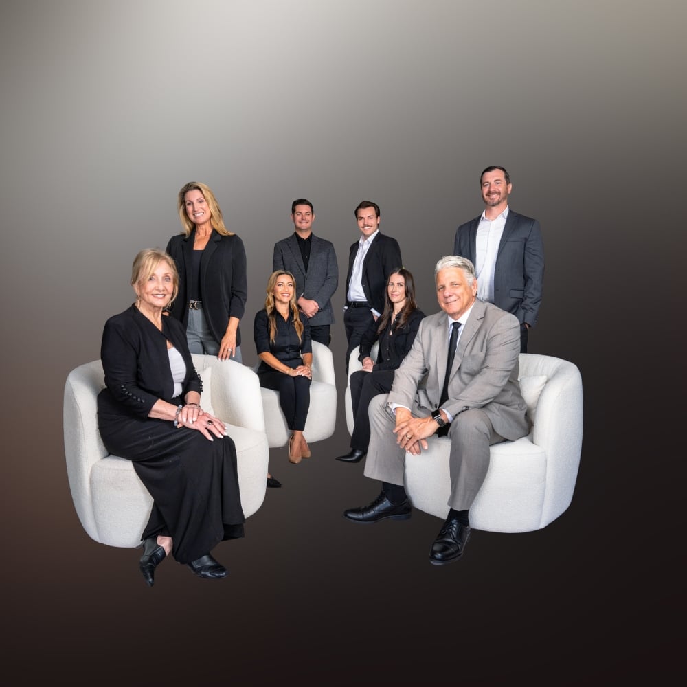 Eight real estate professionals pose together on white chairs against a dark studio background.