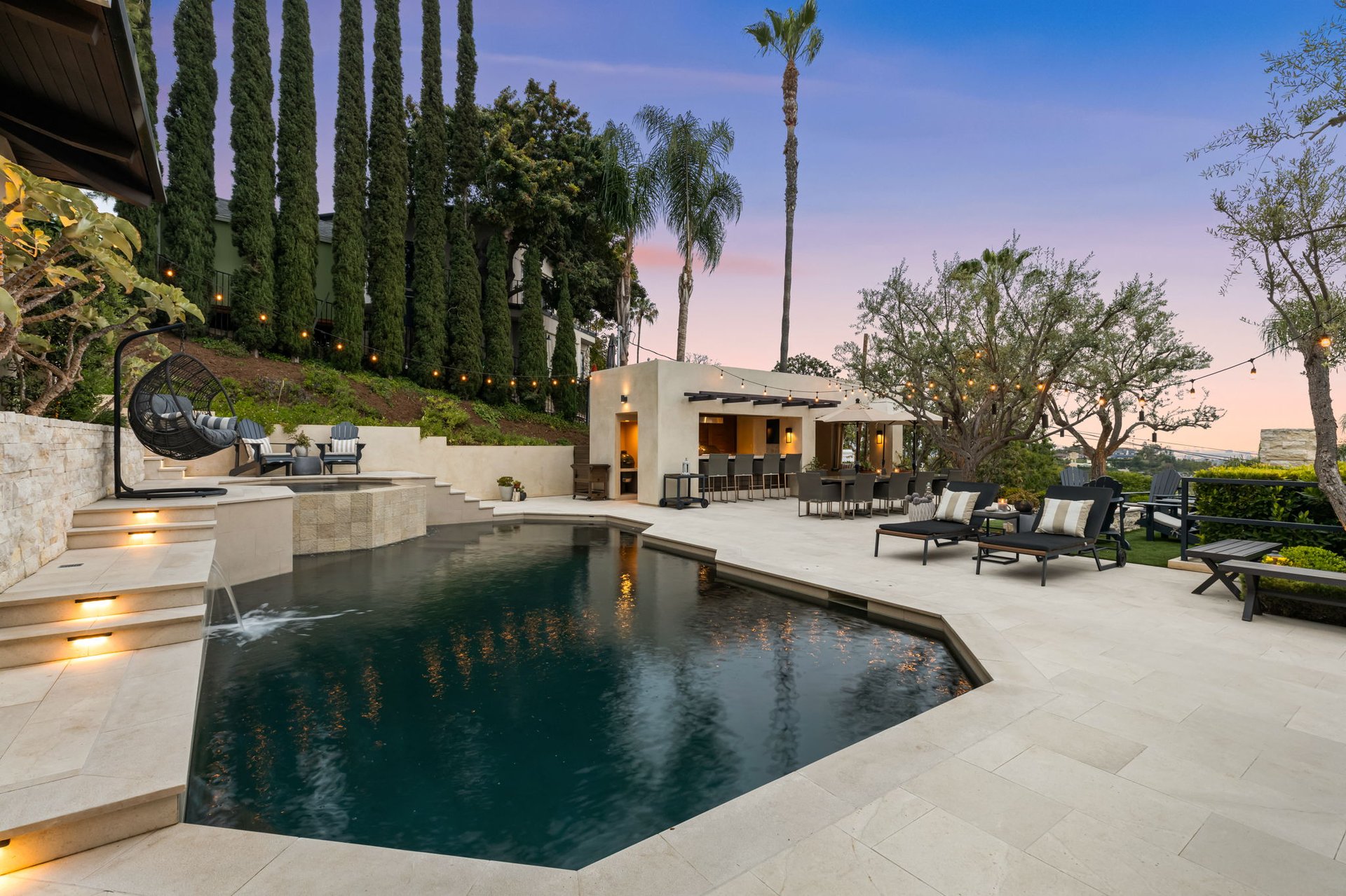 Backyard pool and stone patio with an outdoor kitchen and lounge seating at dusk.