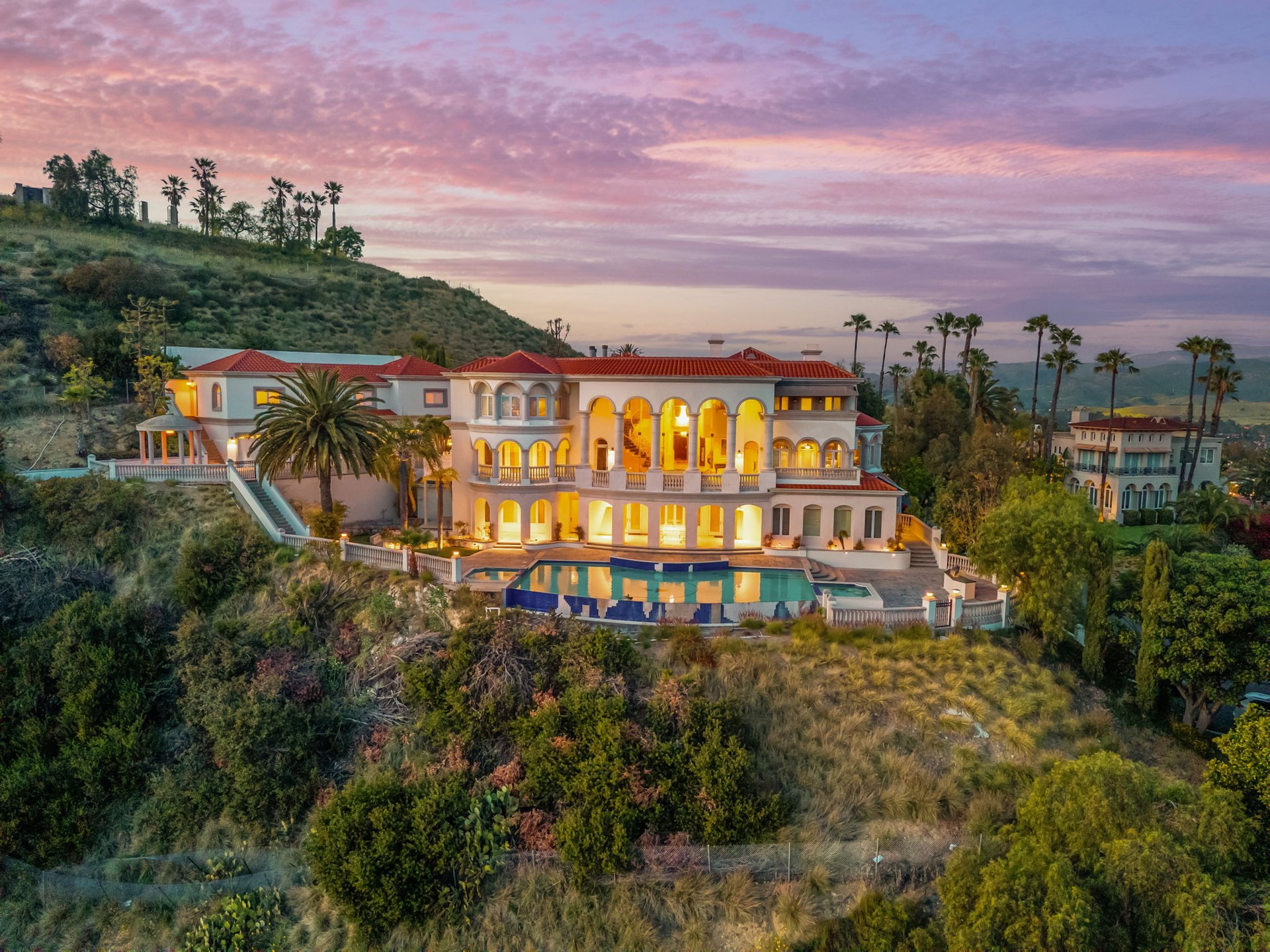 Hillside Mediterranean-style mansion with a swimming pool, palm trees, and a glowing pink sunset sky.