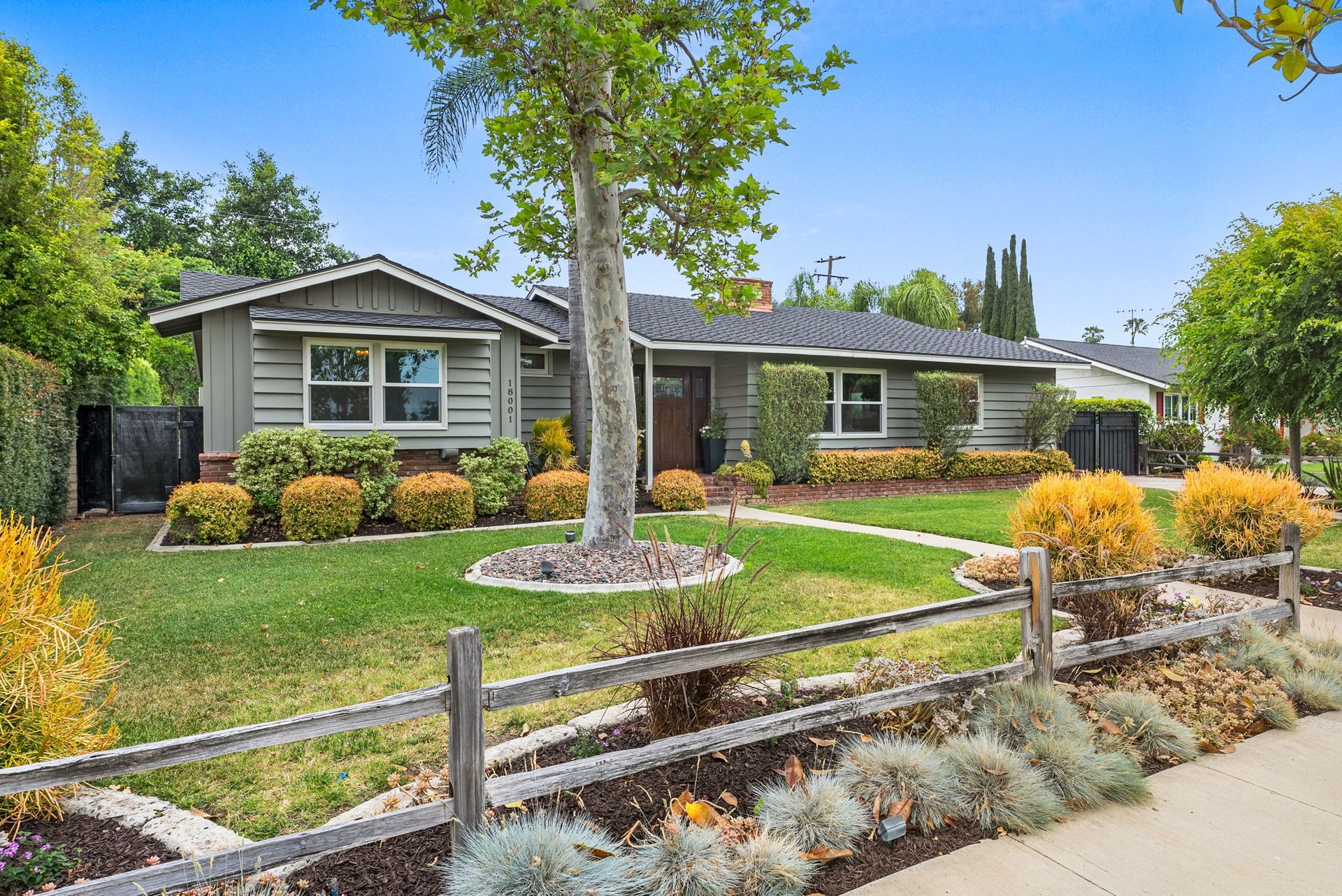 Single-story gray home with a mature front-yard tree, trimmed shrubs, and a low wooden fence.