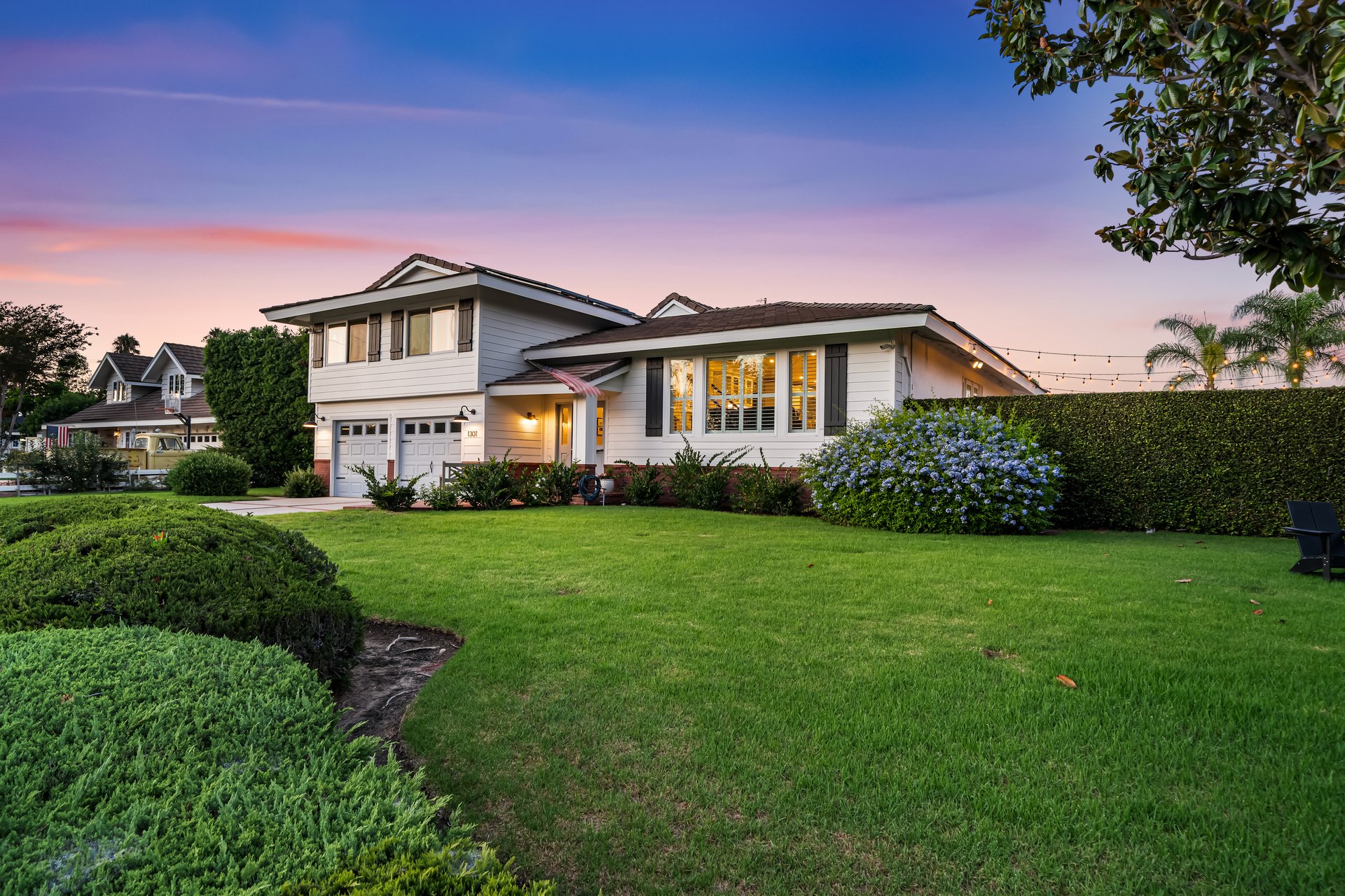 White home with warmly lit windows and a broad green lawn at dusk.