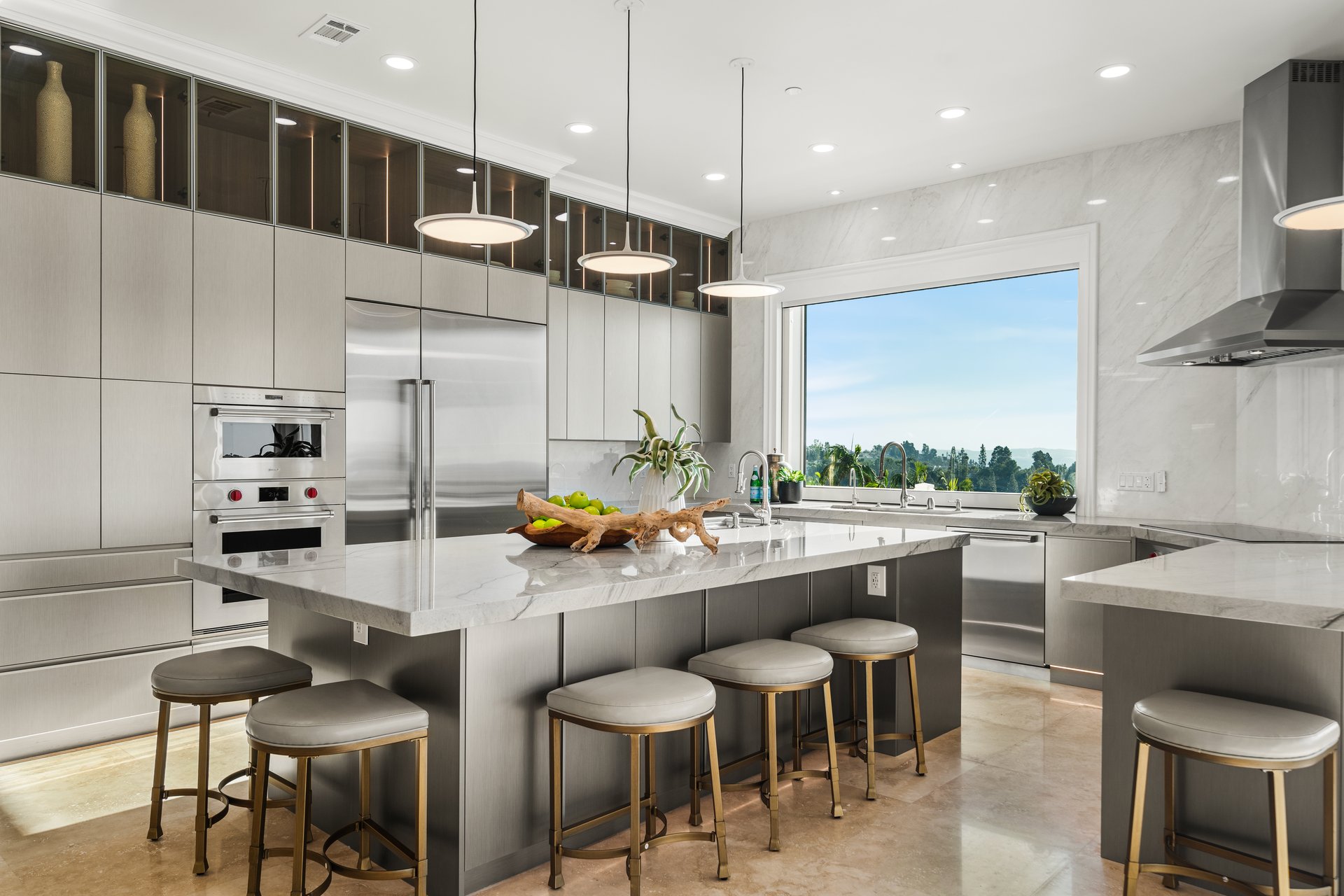 Modern marble kitchen with a large island, stainless appliances, and a wide window overlooking treetops.