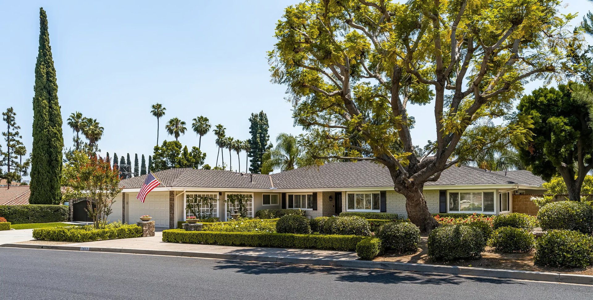 Single-story ranch-style home with manicured hedges, a large shade tree, and an American flag.