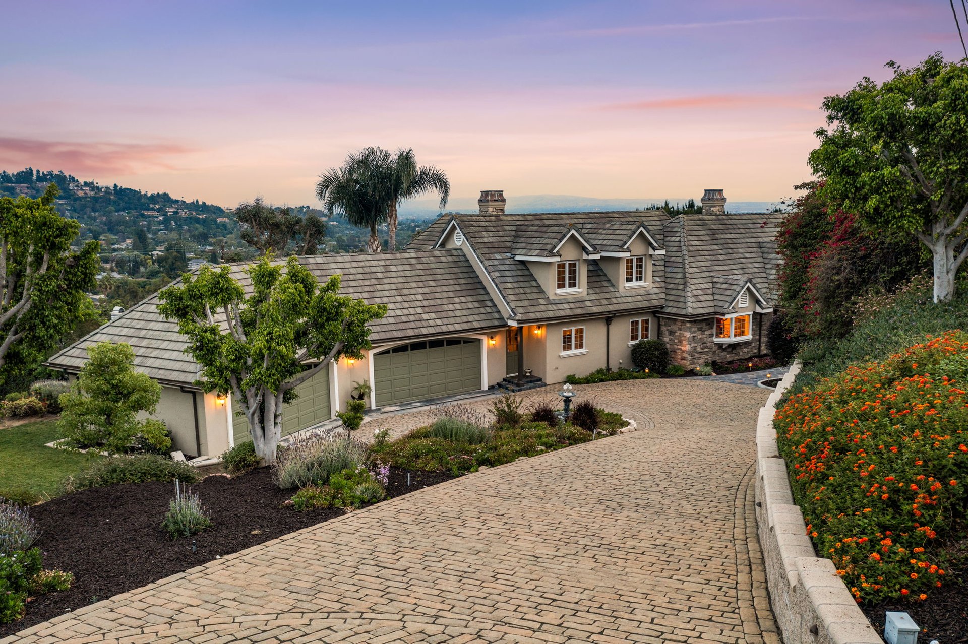 Luxury home with a broad brick driveway, landscaped gardens, and a hillside neighborhood at dusk.
