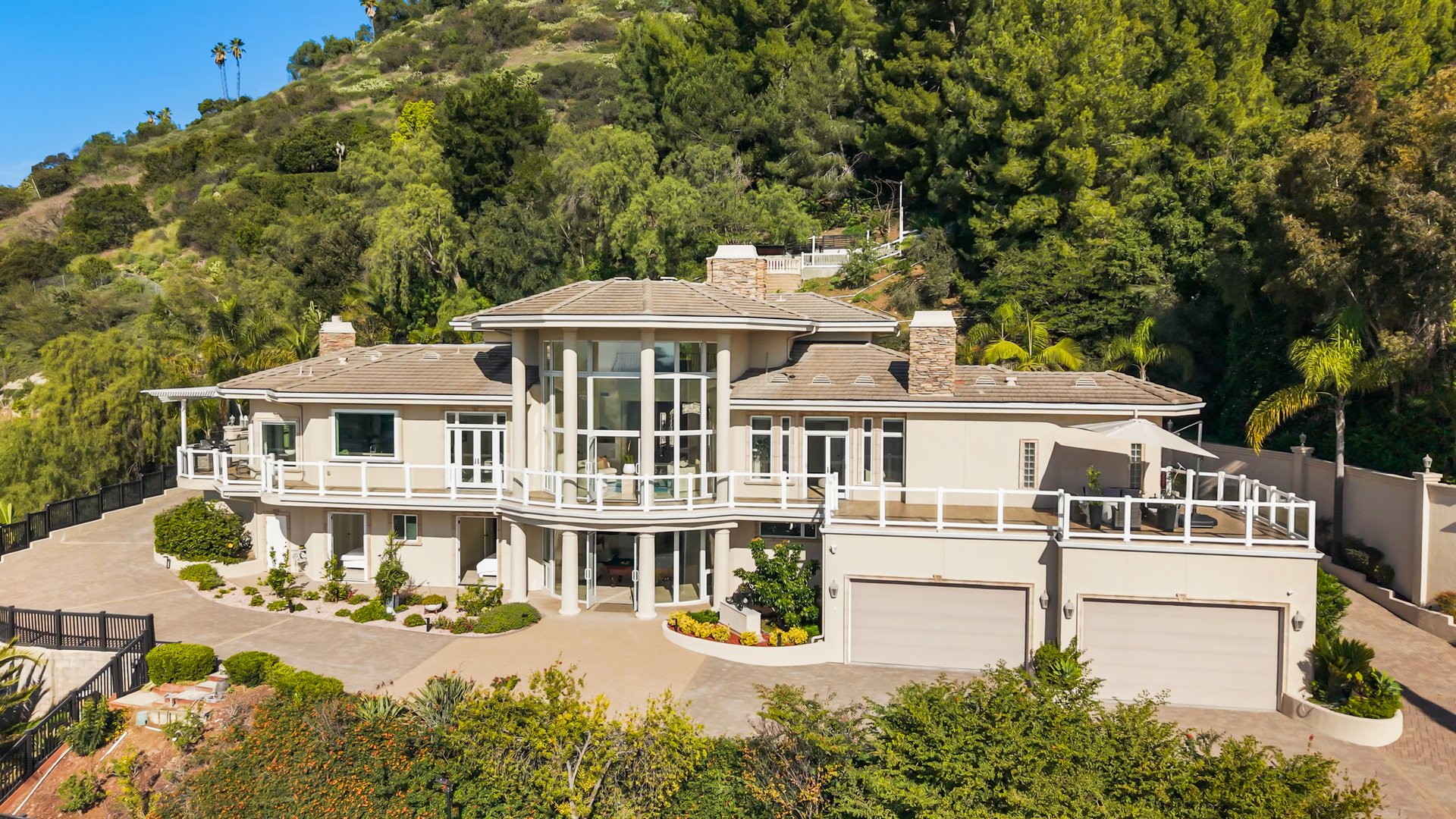 Luxury hillside home with expansive balconies, a three-car garage, and wooded hills behind it.