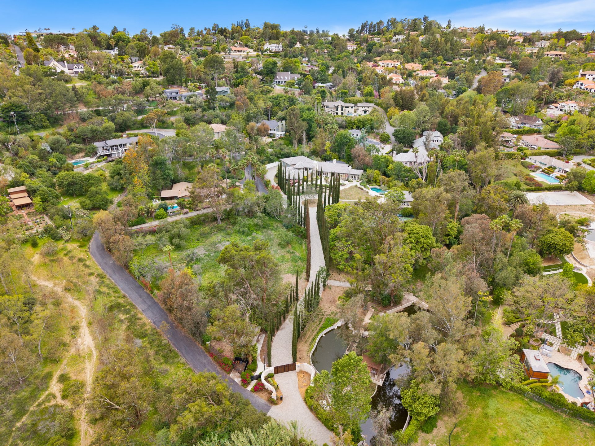 Aerial view of a landscaped estate with a cypress-lined driveway and pond.
