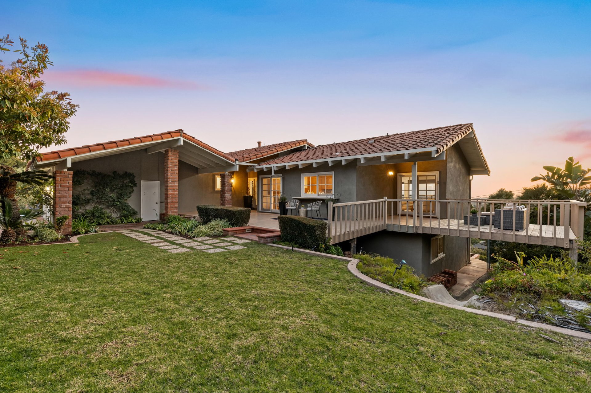 Single-story home with a raised wooden deck and landscaped lawn at dusk.