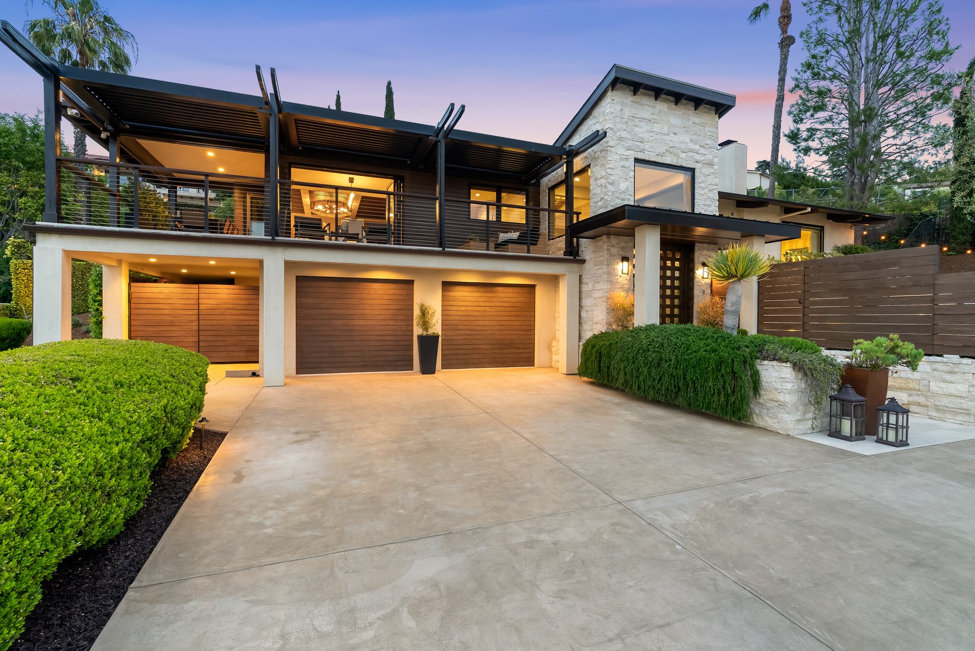 Modern stone-and-wood home with a broad driveway and three garage doors at dusk.