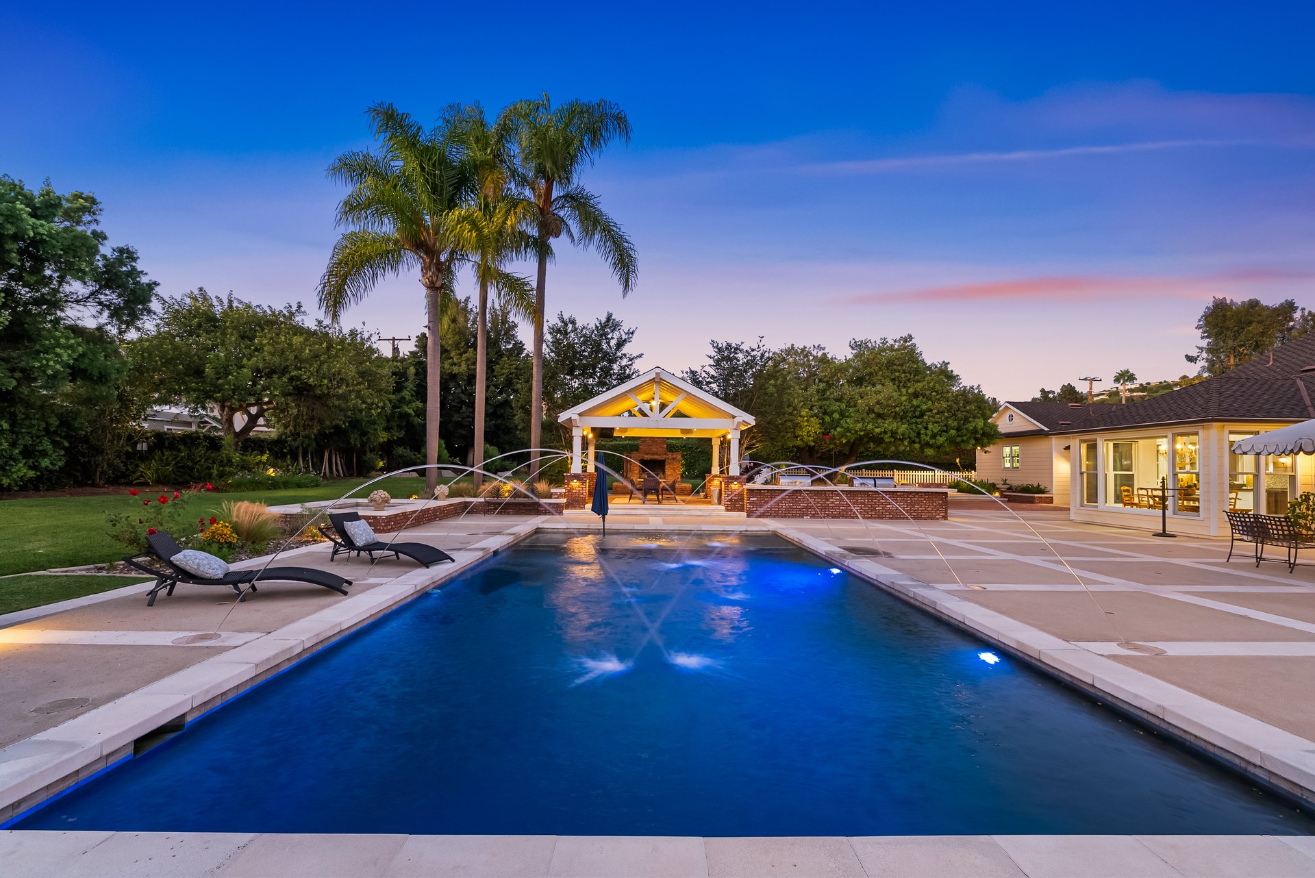 Backyard swimming pool with arcing water jets, lounge chairs, and a lit pavilion at dusk.