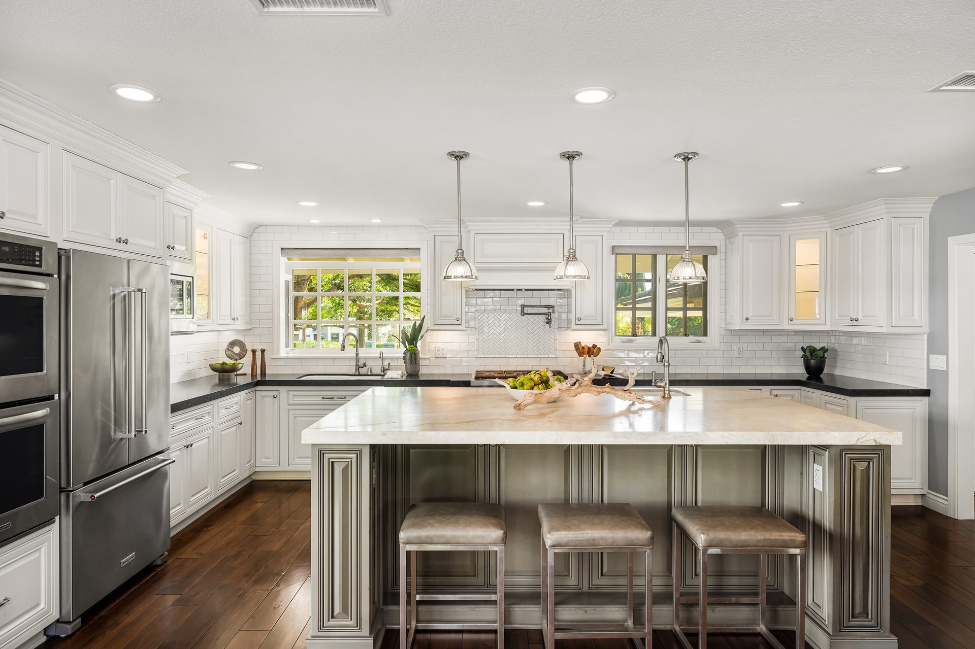 Bright white kitchen with a large marble island, three stools, and windows overlooking greenery.