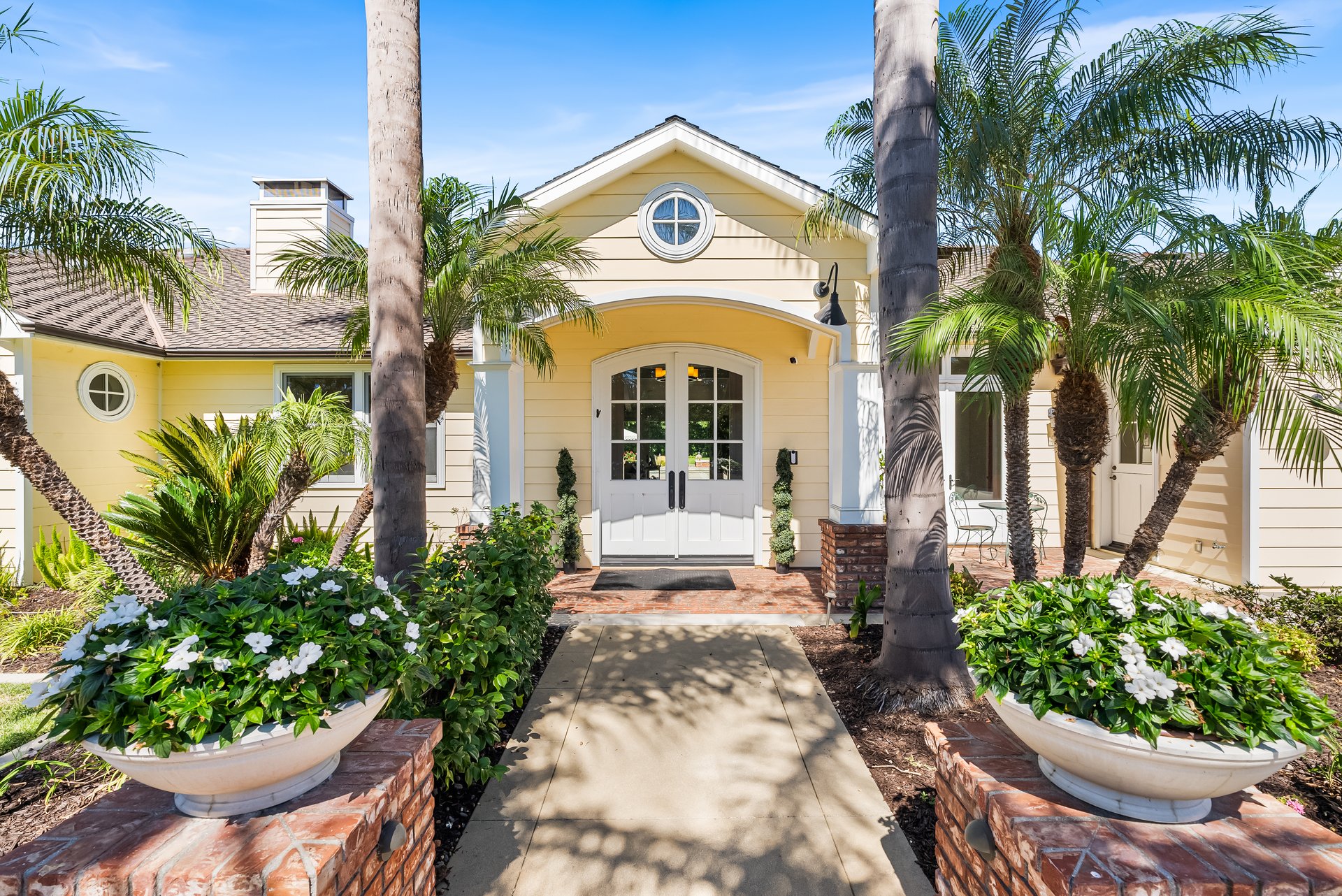Yellow home entrance with white double doors, round windows, palm trees, and flower-filled planters.